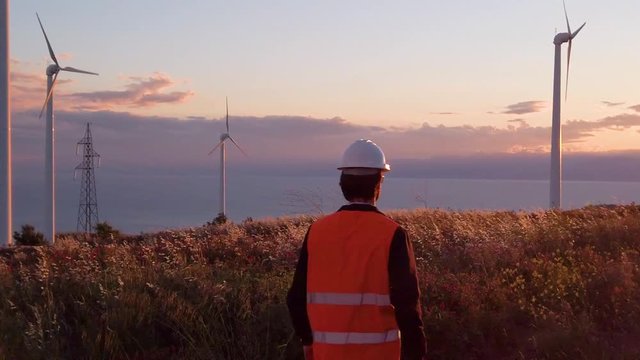 Ingegnere con elmetto e gilet catarifrangente cammina in una parco eolico con turbine del vento. Generatori di energia pulita.