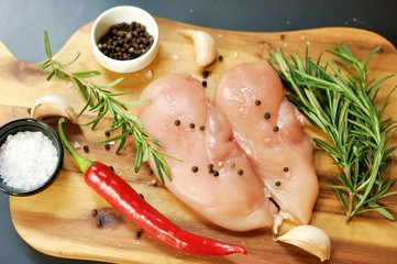Raw fresh uncooked chicken breast meat fillet dish with rosemary, pepper, salt, red chilly pepper and garlic on wooden board and black background. Top dish