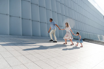 Young family with little daughter walking near the modern building in high-tech architecture style. Man is leading his family. Family holding hands and going ahead. New couple and daughter