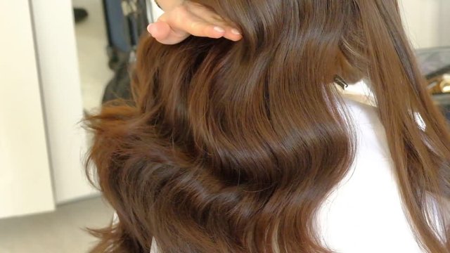 woman hairdresser marcels long brown hair with curling iron in light hairdressing salon backside view