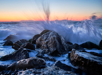waves hitting against the large stones and splashing water high up, sunset in the background