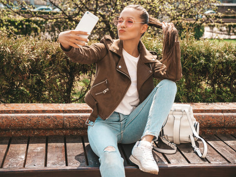 beautiful smiling brunette girl in summer hipster jacket and jeans.Model taking selfie on smartphone.Woman making photos in warm sunny day in the street. Sitting on the bench in sunglasses
