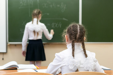 Fototapeta A schoolgirl girl looks into the camera while her friend, a classmate, answers the teacher at the blackboard. obraz