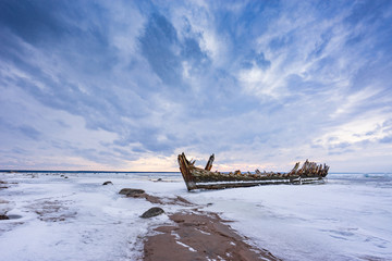 Old broken boat wreck and rocky beach in wintertime. Frozen sea, evening light and icy weather on...
