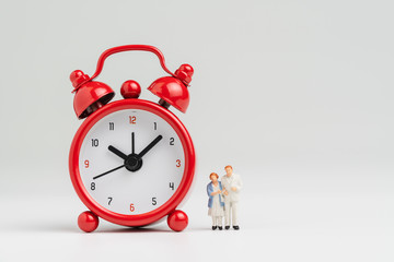 Time counting down for retirement concept, miniature happy senior old couple standing with big red alarm clock on white background with copy space