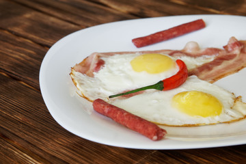 Small sausages, scrambled eggs with bacon and red chilli are in a white plate on a natural wooden surface of pine boards