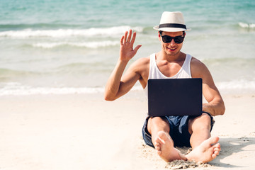 Smiling handsome man relax in sunglasses and straw hat working on his laptop computer on the tropical beach.Summer vacations