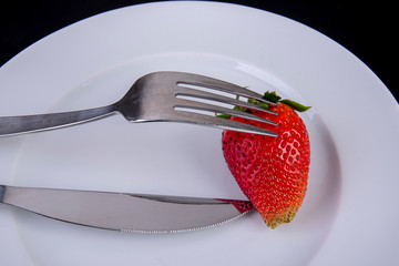 Strawberry is skewered on a fork on a white plate with knife
