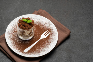 Classic tiramisu dessert in a glass on plate with fork silhouette on dark concrete background