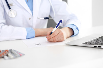 Female doctor filling up prescription form while sitting at the desk in hospital closeup. ...