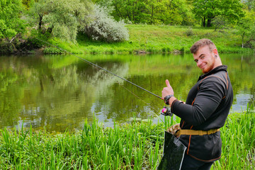 Happy fisherman with a beard on the river bank with a fishing rod in his hands, showing thumbs up