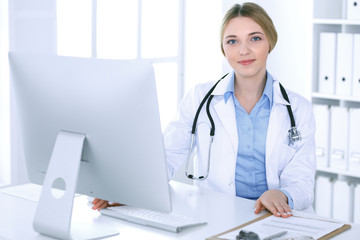 Young woman doctor at work in hospital looking at desktop pc monitor. Physician controls medication history records and exam results. Medicine and healthcare concept
