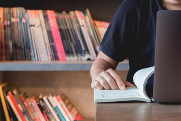 Young business man learning stock trading. Man sitting in library reading book finger pointing, studying book and connecting to internet using laptop making research.