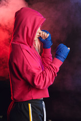 Long-haired blonde kickboxing woman in pink sweatshirt with hood on, training in fitness studio....