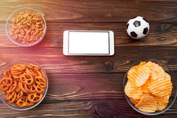 Smartphone with white screen, chips, crackers, snacks, soccer ball on wooden table background. football match. sport. fans.