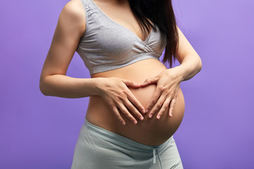 Studio portrait of pregnant caucasian woman with long dark hair. Happy future Mom posing with hands formed heart sign on naked belly over purple background