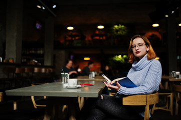 Cheerful young beautiful redhaired woman in glasses using her phone, touchpad and notebook while sitting at her working place on cafe with cup of coffee.