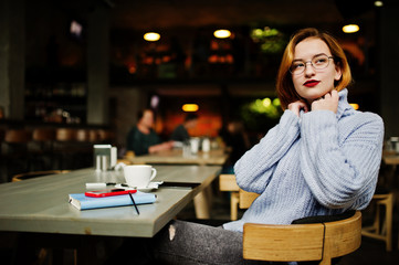 Cheerful young beautiful redhaired woman in glasses sitting at her working place on cafe with cup of coffee.