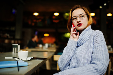 Cheerful young beautiful redhaired woman in glasses sitting at her working place on cafe and speaking by cellphone.