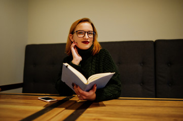 Cheerful young beautiful redhaired woman in glasses, wear on green warm wool sweater,  sitting at cafe and using her notebook.