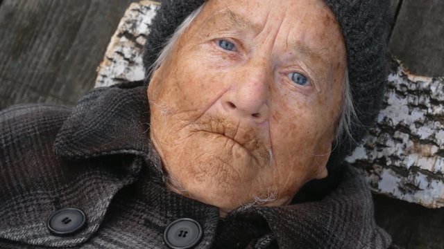 Grandmother lies on the street on wooden bed and looks at camera. Clouse up.  Grandmother is wearing a coat and hat. She looks to the top. It's cold outside.