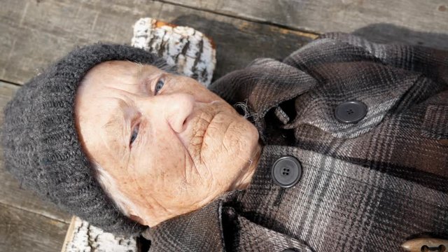 Grandmother lies on the street on wooden bed and looks at sky. Clouse up.  Grandmother is wearing a coat and hat. She looks to the top. It's cold outside.