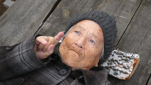 Grandmother lies on the street on wooden bed and looks at sky. Clouse up.  Grandmother looks to the top, then points to up. She is wearing a coat and hat. It's cold outside.
