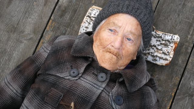 Grandmother lies on the street on wooden bed and looks at sky. Clouse up.  Grandmother is wearing a coat and hat. She looks to the top. It's cold outside.