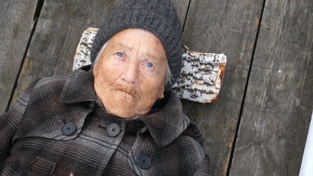 Grandmother lies on the street on wooden bed and looks at sky. Clouse up.  Pan from rotten apples on an old woman. Grandmother is wearing a coat and hat. She looks to the top. It's cold outside.