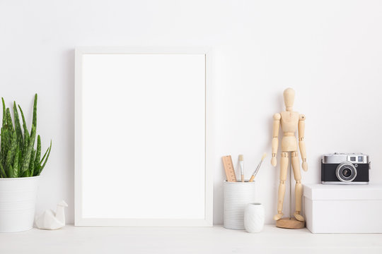 Modern home decor mock up. Creative desk with blank picture frame or poster, wooden mannequin, camera desk objects, artist supplies and plant on a white background.