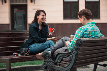 laughing talking man with woman sitting on bench drinking coffee surfing in internet. urban lifestyle
