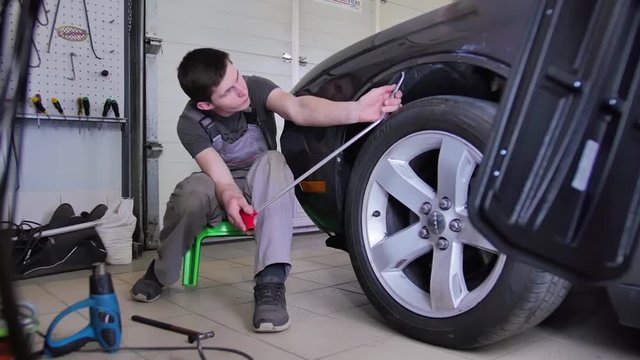 Paintless dent removal. A professional sits on a small stool and repairs a dent in a car without painting with a steel hook. Car repair shop, guy, technology, repair.
