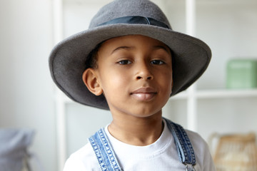 Style, beauty, children’s wear and fashion concept. Close up picture of adorable fashionable eight year old Afro American boy posing indoors wearing stylish round men's hat and denim jumpsuit