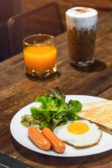 Fototapeta premium Delicious breakfast; fried egg, sausage, toast, salad with orange juice and iced coffee on wooden table.