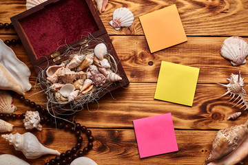 Sea shells in the opened small wooden box or casket on the wooden background surrounded by other sea shells, with chaplet and bright coloured empty stickers. Top view. 