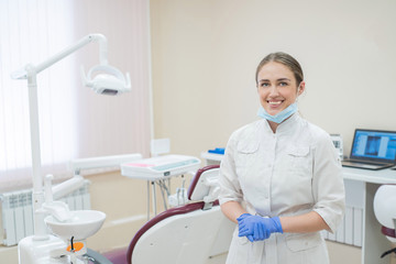 Attractive smiling female doctor in a white uniform at the workplace. A young beautiful female dentist in mask and gloves is standing in the office by the chair