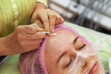 A young girl is lying on a couch during cosmetic procedures with a mask on the face above which beautician woman squeezes body fat and pimples with special metal tool in the form of blade on forehead