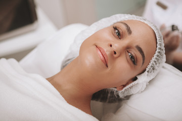 Attractive young woman smiling to the camera, lying on procedure table at cosmetology clinic. Happy female client relaxing after facial treatment at beauty salon. Skincare, dermatology concept