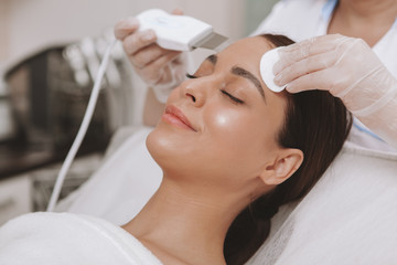Young attractive woman smiling, receiving facial cleanse by professional cosmetologist. Beautician using hardware cosmetology equipment, working with female client. Relaxation, beauty concept