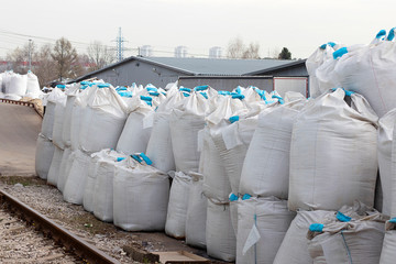 Large white bags with blue ribbon inside the salt lie on the street. Industrial fertilizers are stored in bags in a heap. Near the railway.