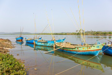 Obraz premium old blue large fishing boats with motors and fishing rods anchored off the coast against the backdrop of a river and green trees on the shore