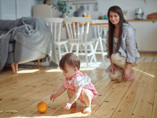 Young mother watching her cute baby daughter playing orange fruit on hardwood floor at home