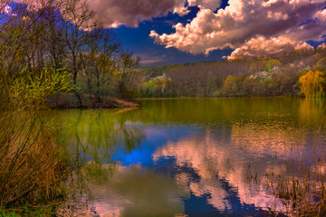 Quiet calm, serene silence over the beautiful mountain forest lake. Reflection of the sky and clouds in the water. The rays of the setting sun beautifully interesting and fascinating illuminate and il