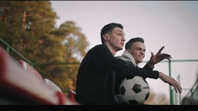 Two young attractive guys sitting with a soccer ball on the chairs of an empty stadium, talking, smiling. The boys watching the sunset, pointing into the distance. Close-up portrait. Slow motion, 4K