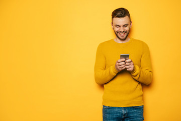 Handsome stylish young beard man in yellow with smart phone in hands is standing over yellow wall