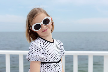 Small (8 years old) pretty cheerful girl in a white dress with black polka dots is standing on a wooden white pier.