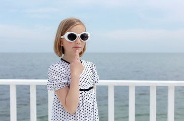 Small (8 years old) pretty cheerful girl in a white dress with black polka dots is standing on a wooden white pier.