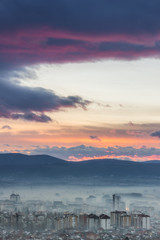 Vertical, soft view of distant city covered by mist and beautiful colors of sunset sky