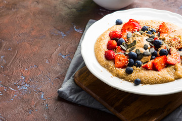 Delicious amaranth topped with almond butter, fresh strawberries and blueberry, sesame, pumpkin seeds. Plant based breakfast served on dark wooden board. Healthy colorful gluten free food concept.