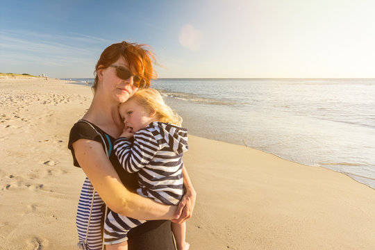 Junge Frau mit Kind auf dem Arm steht entspannt am Strand und genießt den Sonnenuntergang